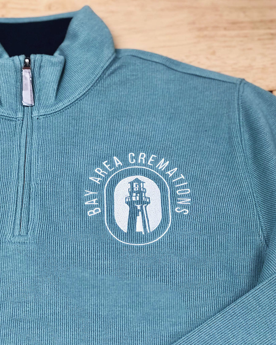 A sage quarter-zip embroidered in white with the Bay Area Cremations lighthouse logo