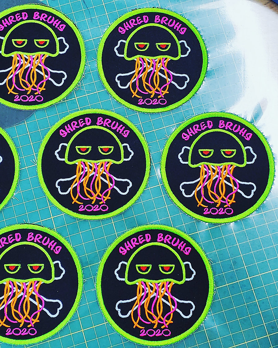 Round Shred Bruhs patches on the cutting mat: a neon squid over crossbones in green, pink and orange thread