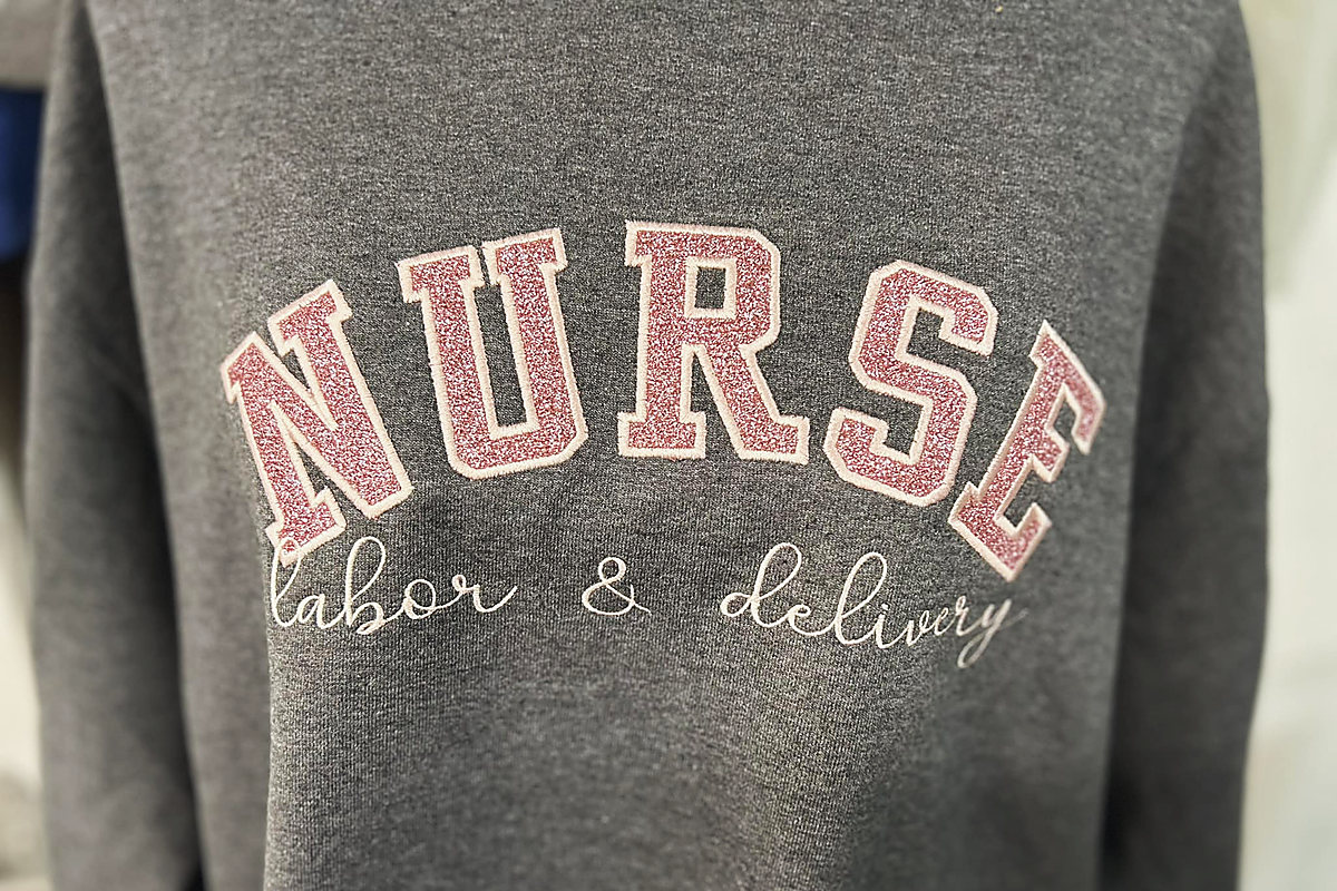 A grey sweatshirt with NURSE in pink glitter appliqué and labor and delivery stitched beneath in script